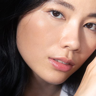 A imagem mostra uma mulher amarela, de cabelo preto com uma das mãos próxima ao rosto. A iluminação destaca a pele uniforme e os lábios hidratados